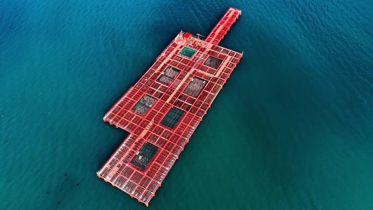 Drone view of red aquaculture platform on teal sea. Elongated fish cage structure with grid frames and net pens surrounded by calm deep green water.