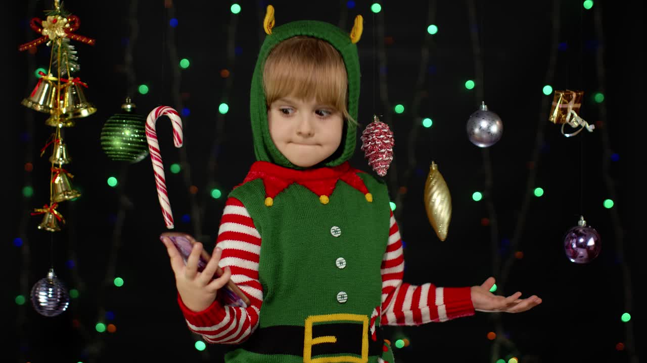 Kid girl in Christmas elf Santa Claus helper costume making selfie on smartphone. New Year holidays