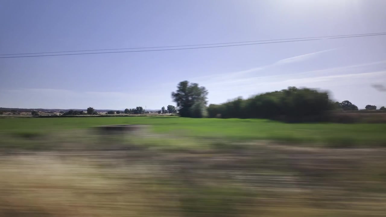 Scenic view from a car window driving quickly through the rural landscape of Lleida, Catalonia, with green and golden fields passing by under a clear blue summer sky in sunny Spain