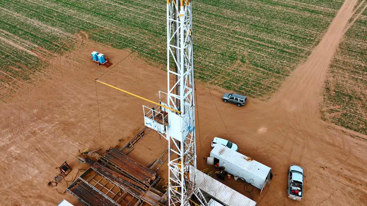 Rising above the drilling site along the high tower for oil boring. Cars and metal pipes are at the location.