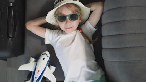 Preschooler-girl-in-sunglasses-looks-in-camera-lying-on-sofa
