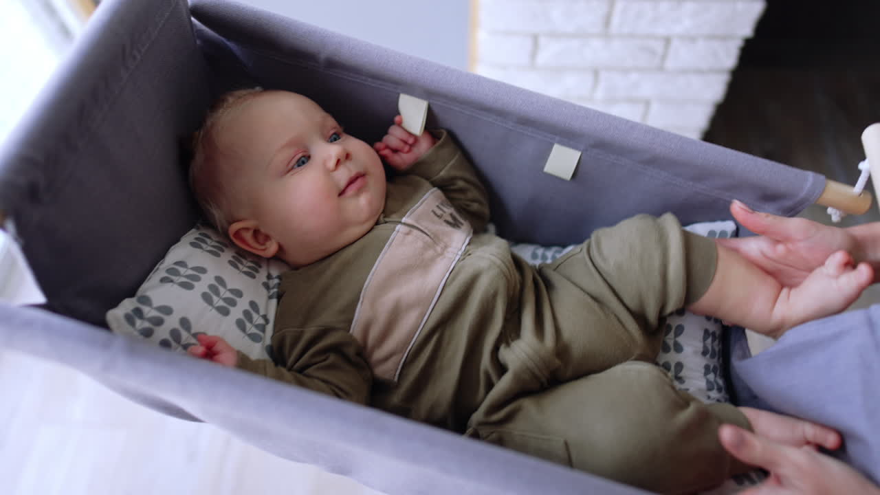 Beautiful Caucasian infant lies in his little cradle. Mom's hands hold baby by the ankles and he smiles.