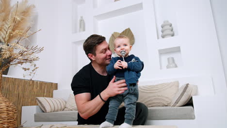 cute little baby held by the father indoors. dad waves goodbye to the camera and kid with pacifier looks ahead. low angle view.