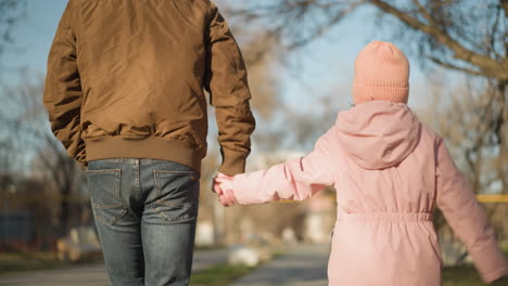 father in brown jacket and black jeans playfully jumps with his daughter in a pink cap and jacket, both enjoying a sunny day in the park