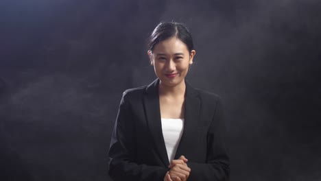 smiling asian speaker woman in business suit clapping her hands and looking around while standing in the black screen studio