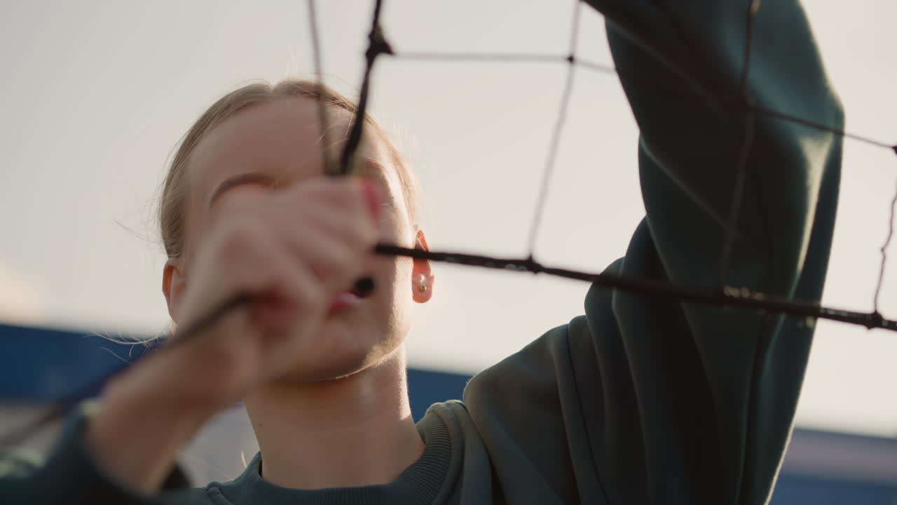 Close-up of volleyball net held by person in green sweater checking its tension with golden light reflecting around them, face slightly blurred