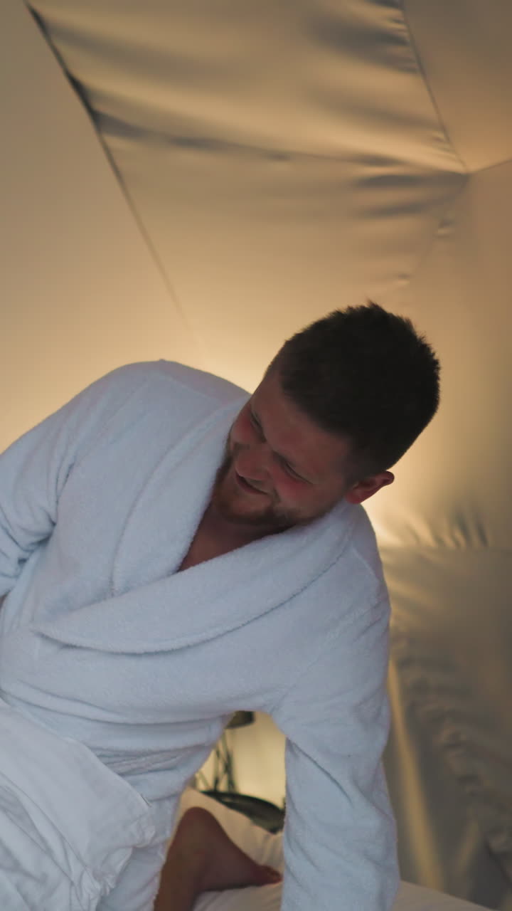 Woman hits man with pillow on bed at glamping. Positive lady in bathrobe plays fighting with boyfriend in round house. Couple fun on honeymoon