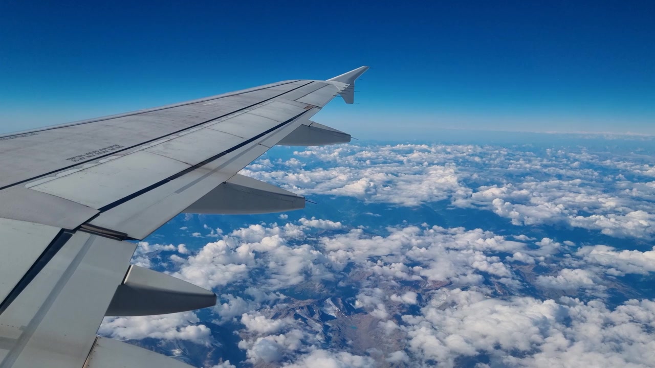 Vista dal finestrino dell'aereo dell'ala che vola alta sopra la Terra, circondata da soffici nuvole bianche sotto un cielo blu infinito, che evoca viaggi aerei, aviazione, libertà e un senso di prospettiva