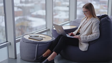 comfortable workplace in fashionable new office premise. lady sitting in a bean bag chair working on laptop. panoramic window backdrop.
