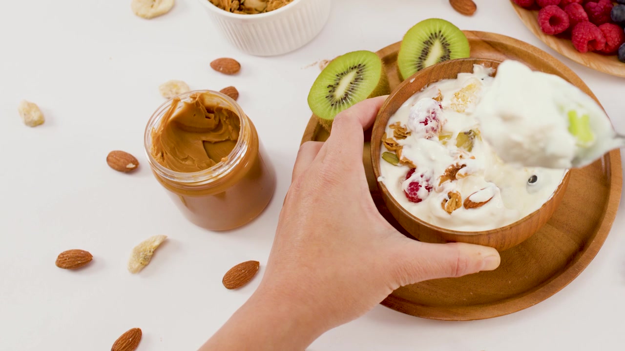 Overhead shot of hands scooping yogurt from a wooden bowl filled with berries and nuts.