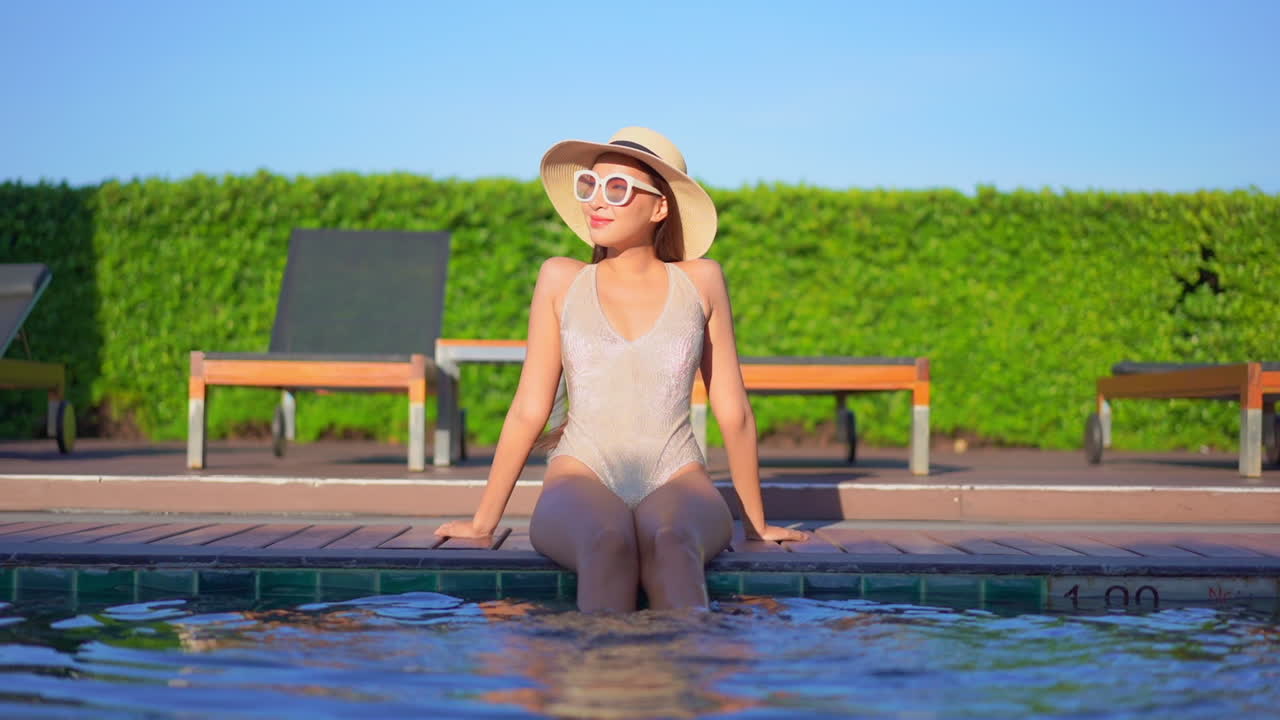 나무 수영장 데크에서 휴식을 취하는 예쁜 젊은 여성이 물속에서 다리를 걷어차면서 태양을 즐깁니다.