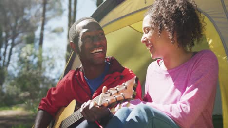 Smiling-diverse-couple-sitting-in-tent-and-playing-guitar-in-countryside