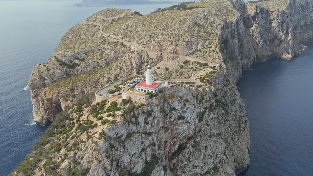 Flying around a white lighthouse with a red roof stands prominently on a dramatic rocky cliff. Winding roads lead up to the structure, surrounded by rugged terrain and sparse vegetation.