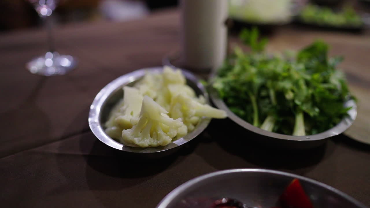 Still camera clip featuring chopped cauliflower and celery on small aluminum bowls over dinner table for traditional cuisine preparation.