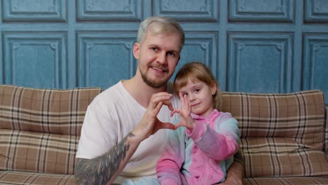 Father-and-child-daughter-kid-in-pajamas-sit-on-couch-smiling,-making-sign-of-shape-heart-near-chest