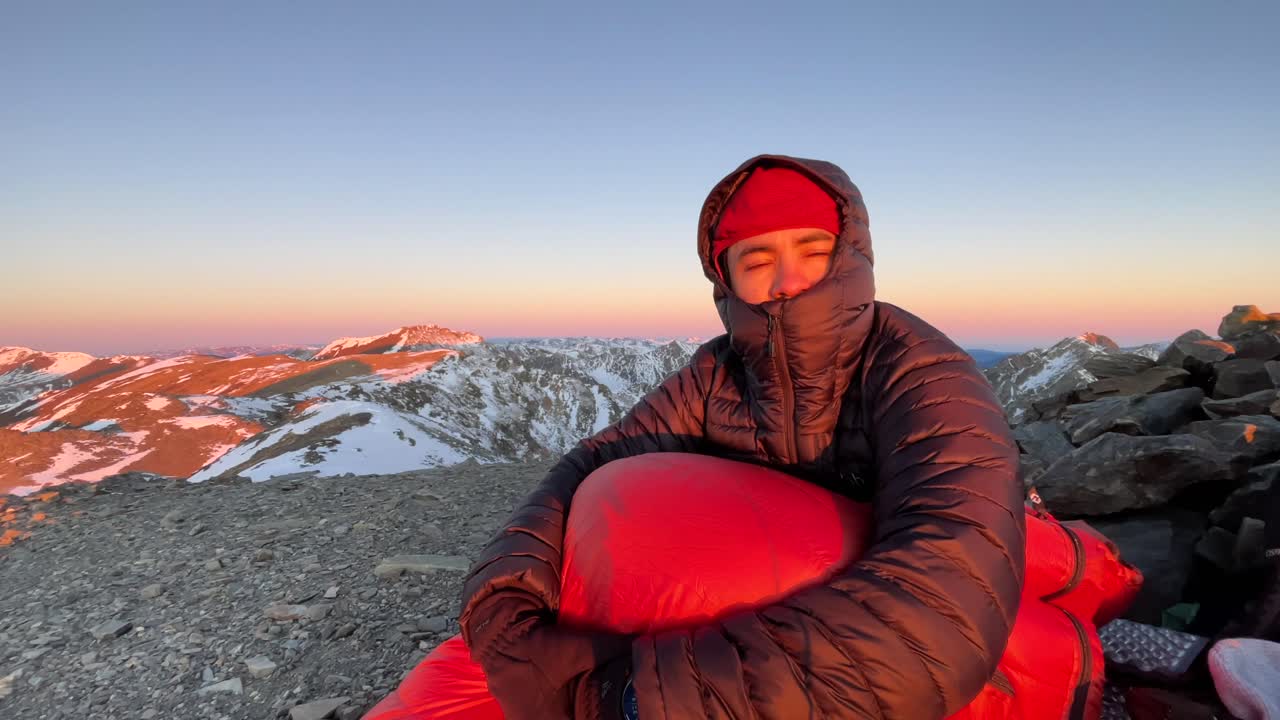 Kayaklıklar giymiş bir dağcı, güneşin doğuşuyla karlı Pireneler zirvelerinin aydınlandığı kayalık bir zirvede uyku tulumunda dinleniyor. Sakin kış atmosferi, soğuk havada kamp, sabahın erken saatlerinde geniş dağ manzarası