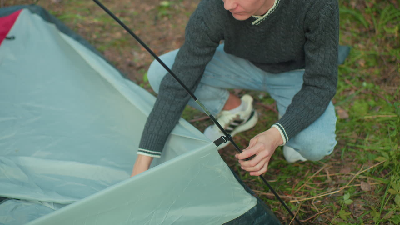 young man in dark sweater crouch on forest ground carefully attach flexible pole to tent corner with both hands focused on setup process surrounded by grass dry leaves and early morning light