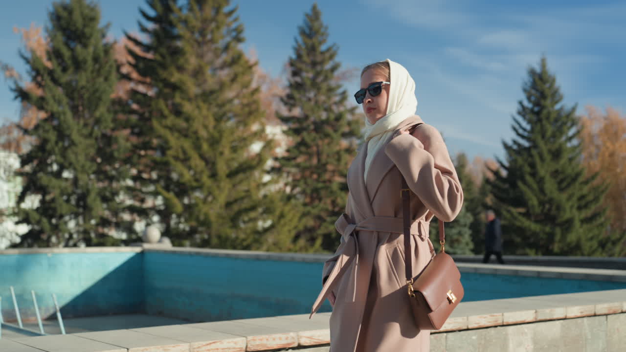 Donna in cappotto beige vicino a una fontana, con foulard bianco e occhiali da sole, cammina attraverso un parco autunnale fiancheggiato da betulle e alberi sempreverdi. Porta una borsa in pelle. La luce soffusa del sole proietta un caldo bagliore.