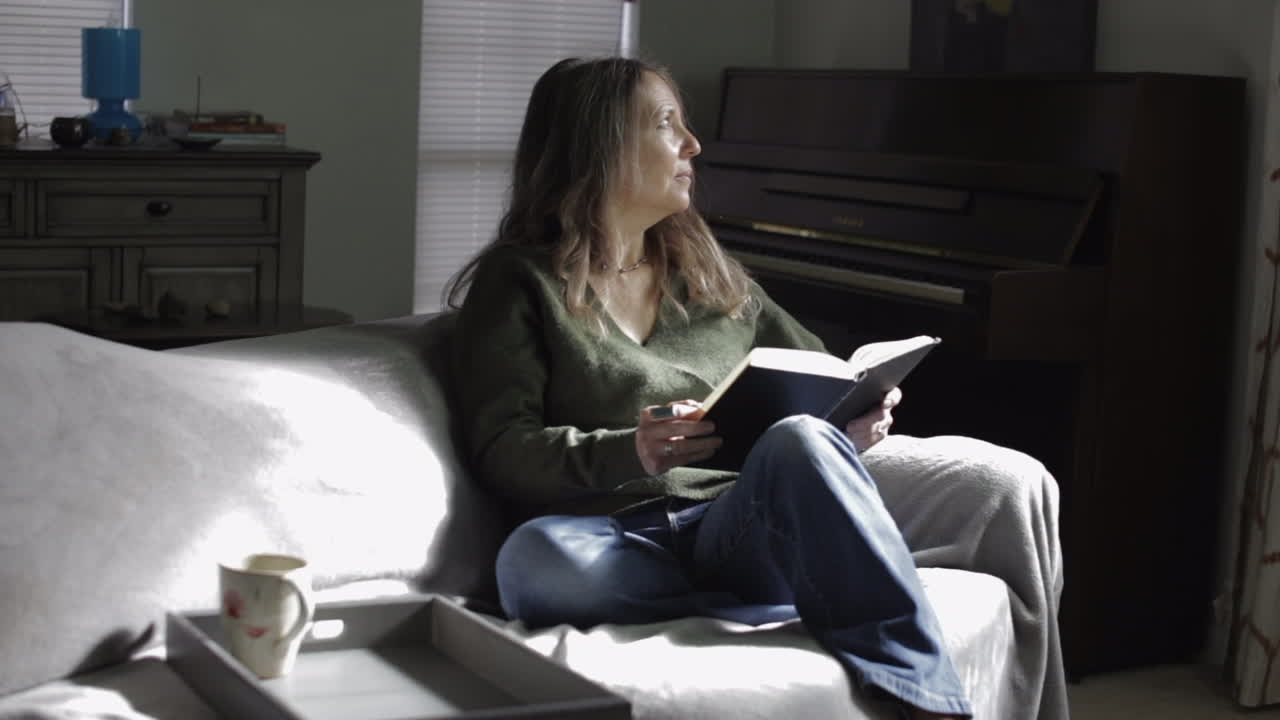 Beautiful Adult Caucasian Woman Pondering Thoughts While Sitting And Holding Book At The Living Room