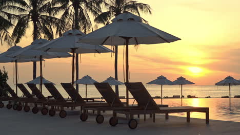 a colorful sunset highlights a line of empty sun loungers and beach umbrellas along the edge of a resort swimming pool