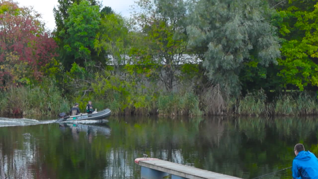Small motorboat with an engine riding or driving down a narrow small river that is reflecting the surrounding trees and plants during summer in slow motion. Boat creates waves and ripples in the water