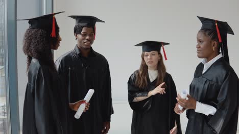 two young women and one young african-american man and one young caucasian female university graduate with diplomas and books in their hands stand by the window and talk.