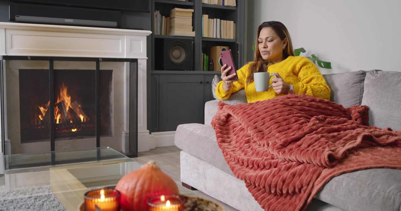 Mujer afroamericana en casa, con enfoque selectivo, deslizando un teléfono rosa, leyendo mensajes y bebiendo de una taza.