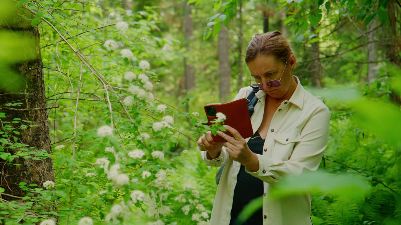 Una donna nella foresta esamina una pianta con un taccuino. Indossa occhiali e una giacca leggera. Il verde la circonda con fiori bianchi. La scena cattura uno studio della natura o una ricerca botanica. Ideale per scopi educativi.