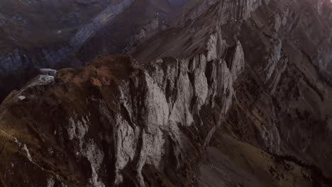 Aerial-flyover-over-Schaefler-in-Appenzell,-Switzerland-at-sunset-with-pan-up-motion-from-the-mountain-hut-above-the-cliffs-towards-Santis