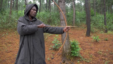 a very slow motion shot of an african hunter wearing a cloak in a forest taking aim with a bow and arrow and shooting