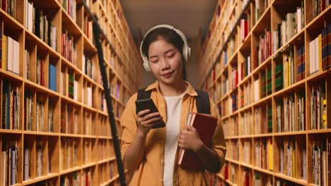 una estudiante asiática sonriente con una mochila y algunos libros con auriculares y usando un teléfono inteligente mientras está de pie en la biblioteca