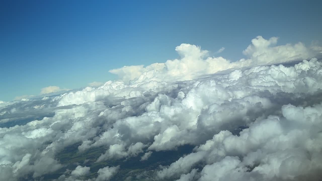 An aerial cloudscape recorded from a jet cockpit while flying at high altitude over a layer of cotonny clouds, with threatening storm clouds under a blue sky bathed with the sunset light.