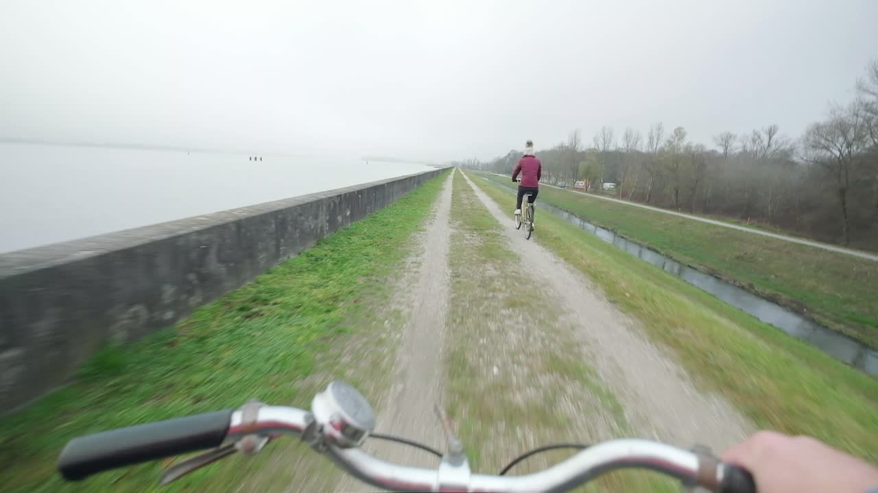 Following a lady riding a bike next to a lake on a overcast day. Handle bars of another bike in the foreground as looks onto another lady riding a bike on a bike track in Slovenia