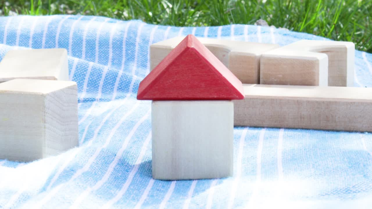 집 보험 개념: 한 손이 나무 에코 큐브 장난감에서 집에 빨간 지붕을 고 있습니다. 공원, 자연 배경에서 담요에 장난감. 은 봄 날.