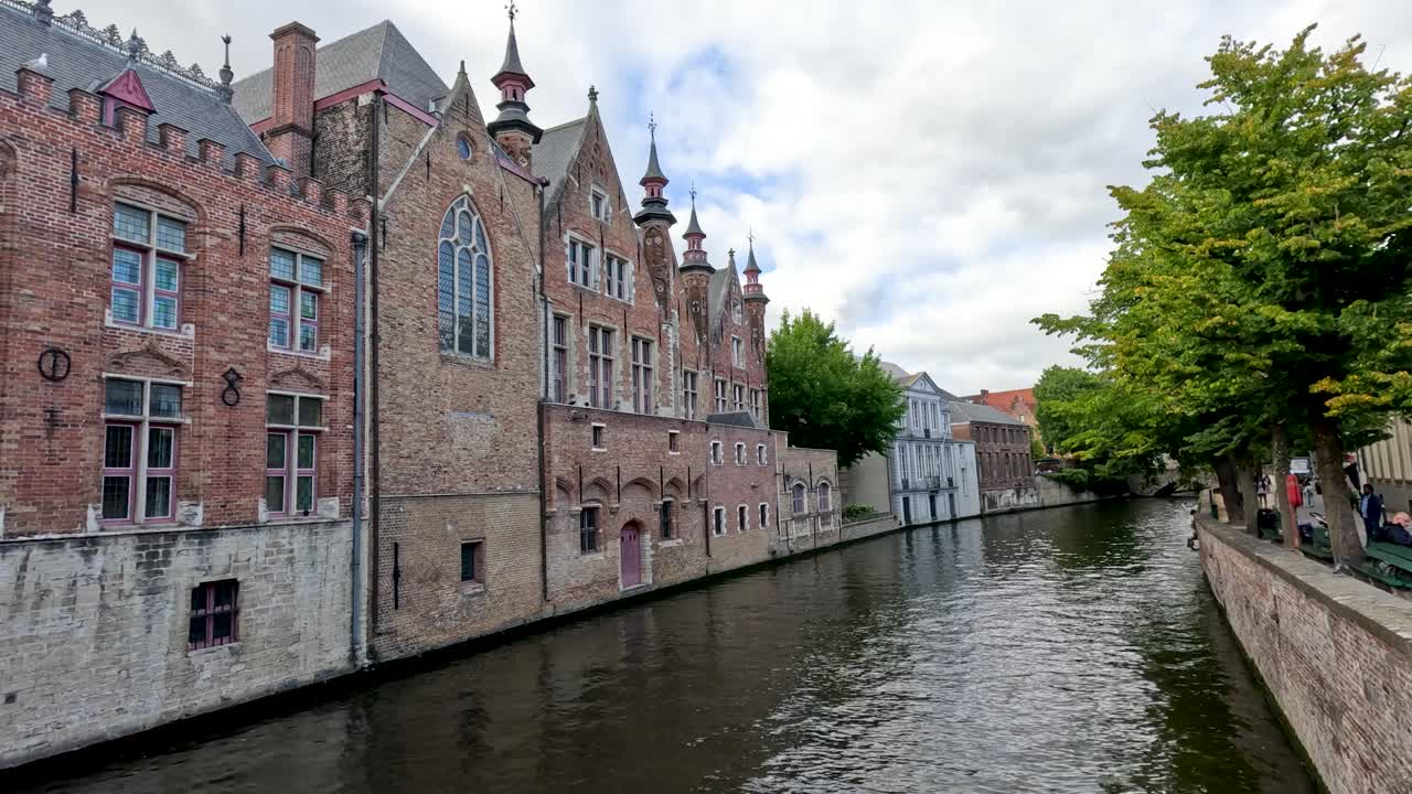 Camera glides along Bruges canal, showcasing medieval brick architecture, calm water, and lush summer trees