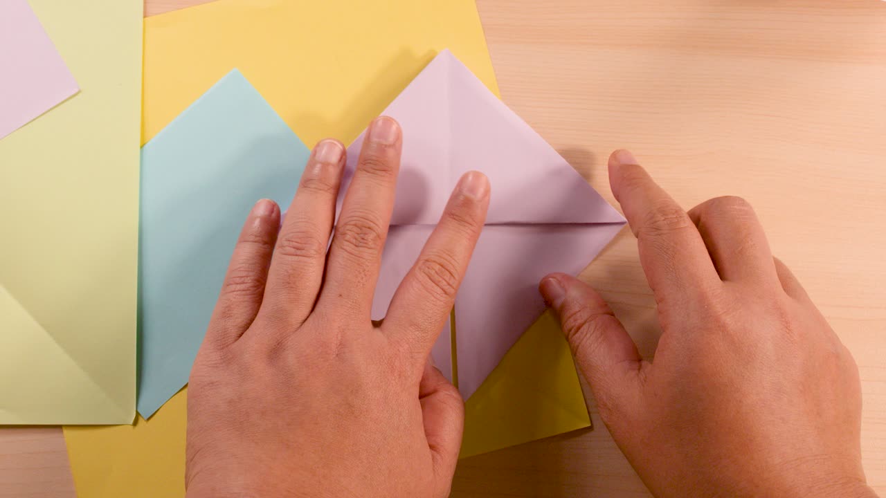 Person folds pastel origami paper on desk under soft lighting, top-down camera, step-by-step process