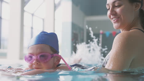 Female-Swimming-Coach-Giving-Girl-Holding-Float-Lesson-In-Pool