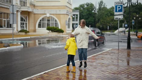 A-happy-woman-in-a-white-jacket-together-with-her-teenage-daughter-in-a-yellow-jacket-are-standing-on-the-edge-of-the-road-and-they-are-doused-with-water-by-a-car-driving-through-a-puddle-after-the-rain