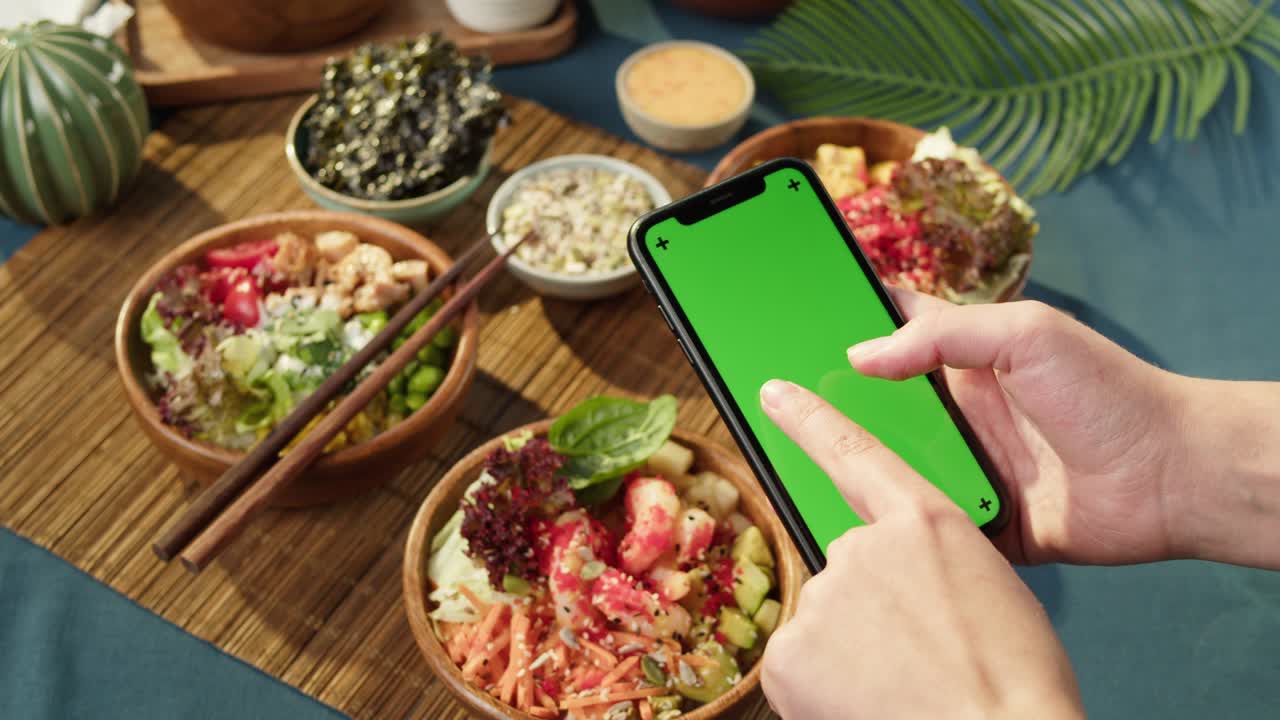 tazones de poke hawaianos, sosteniendo un teléfono inteligente con pantalla verde cromada de primer plano. poke cocido hecho de verduras en rodajas, mariscos y verdura. platos vegetarianos saludables. comida cruda vegana asiática, palillos