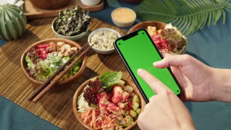 tazones de poke hawaianos, sosteniendo un teléfono inteligente con pantalla verde cromada de primer plano. poke cocido hecho de verduras en rodajas, mariscos y verdura. platos vegetarianos saludables. comida cruda vegana asiática, palillos