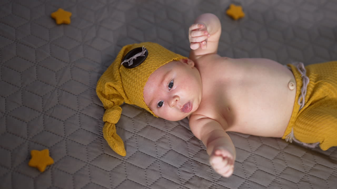 Cute child in a yellow cap and pants. Baby with naked tummy lying on the bed. Little toy stars around the child.