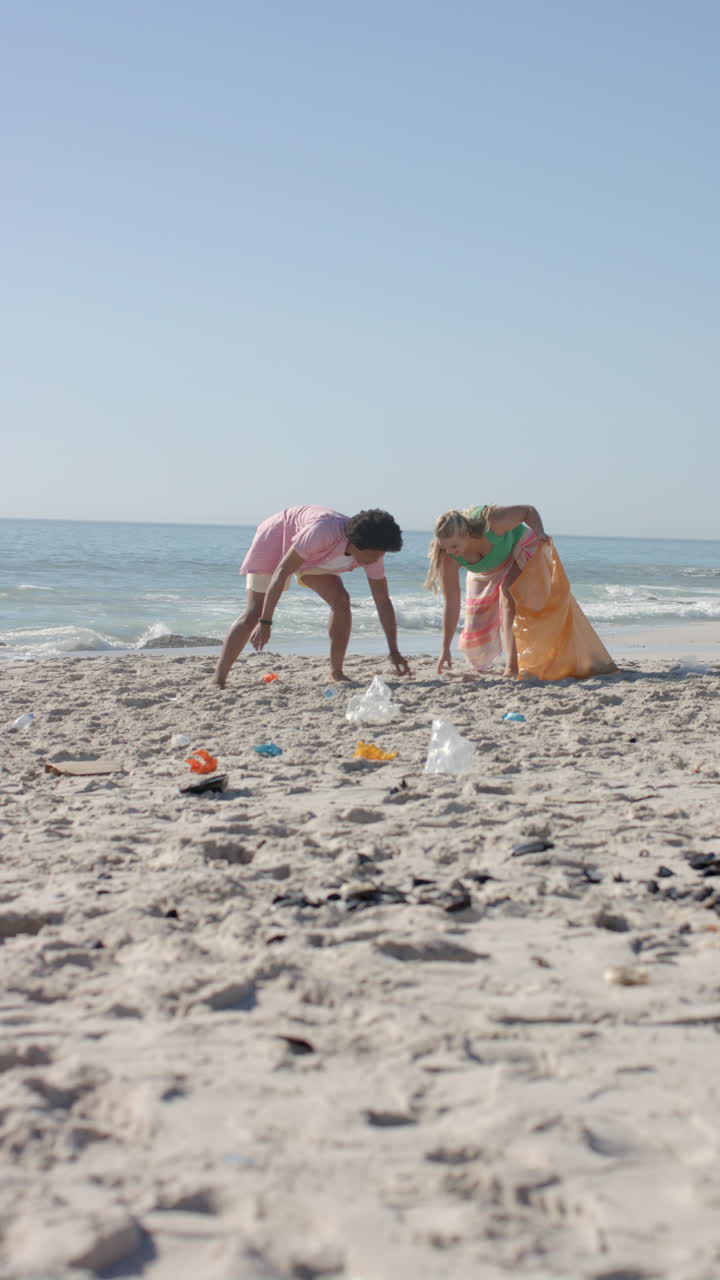 Vertical video: Cleaning beach, group of friends picking up trash from sand near ocean