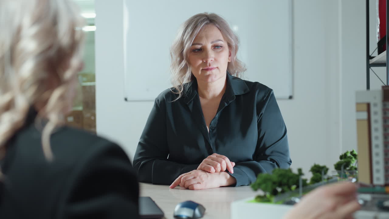 rear view of real estate professional explaining project to client seated across desk with detailed building miniature on table bright office setting suggests business interaction