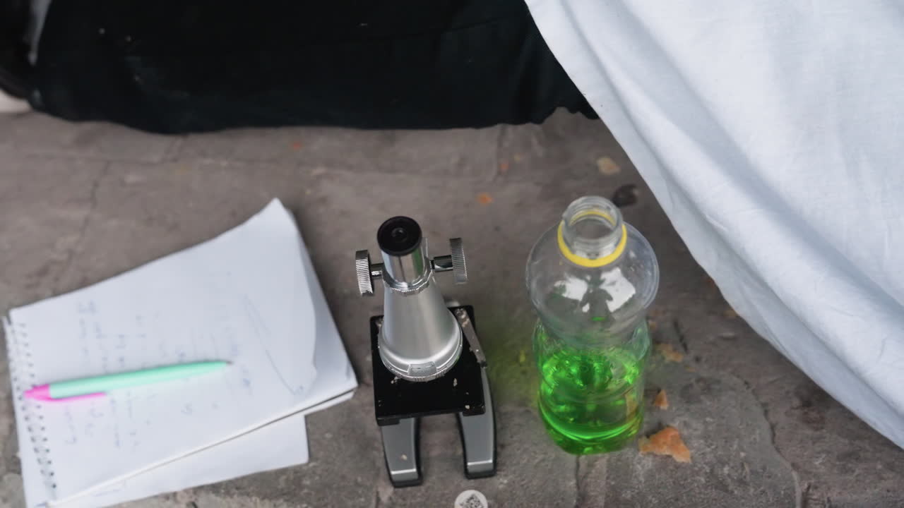 Close view of microscope placed on ground beside bottle filled with green liquid and notebook with pen, reflecting atmosphere of scientific study, experiment, or research activity captured