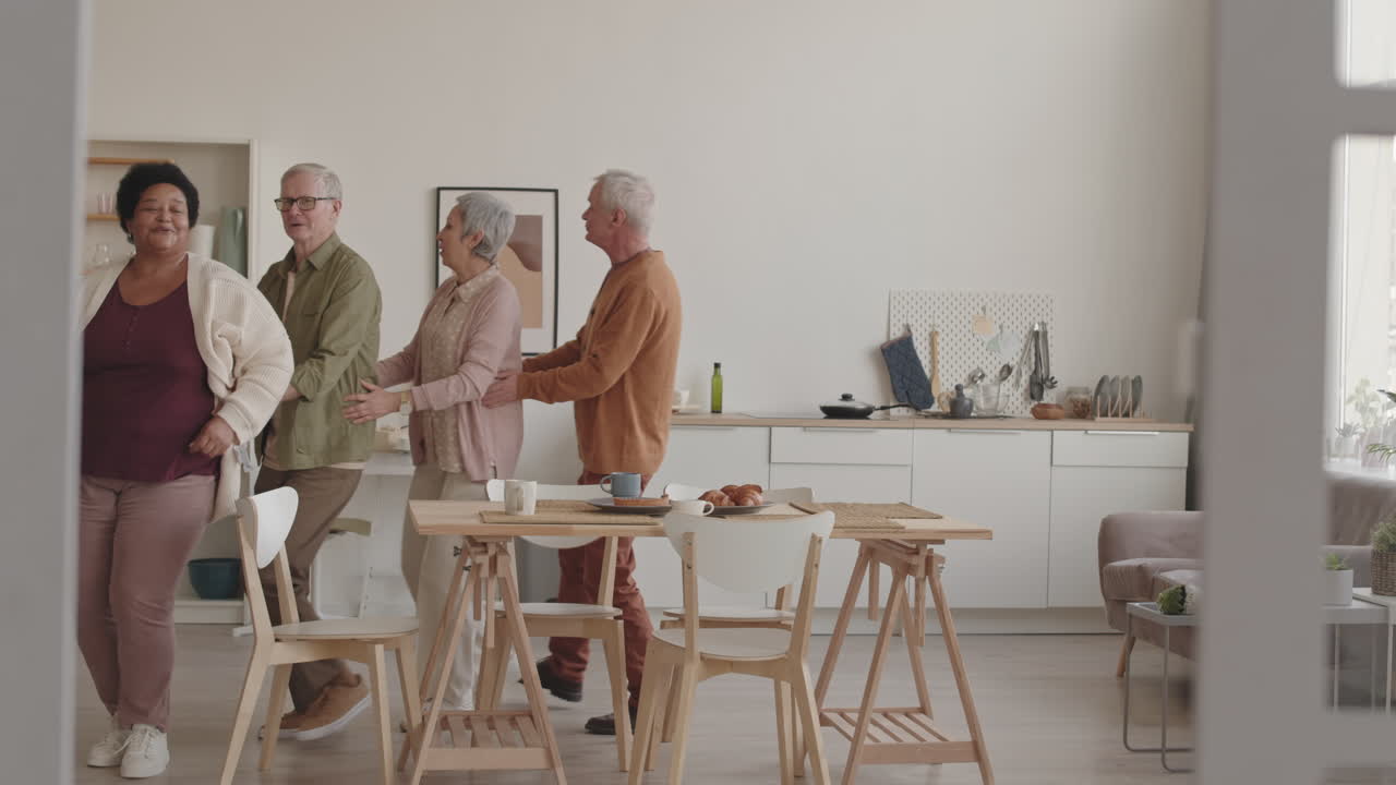 Slow motion wide of multiethnic senior friends dancing conga at home around dining table