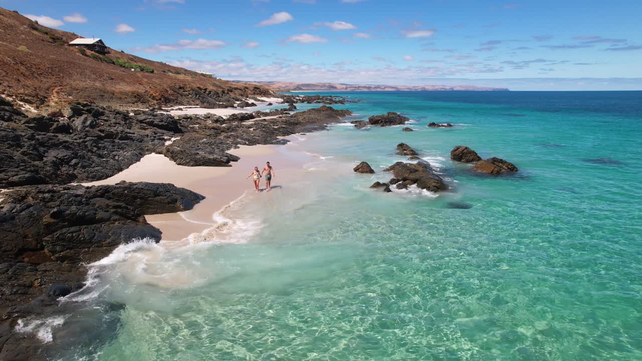 una pareja camina de la mano en una playa de arena blanca en el océano azul brillante y cristalino y comienza a nadar y jugar mientras no hay nadie más a la vista