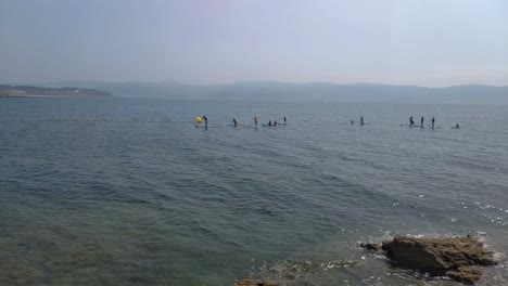 Gente-En-Un-Grupo-Tomando-Clases-De-Paddle-Surf-Con-Bollos-En-Un-Día-Soleado-Al-Amanecer-En-La-Costa-Con-El-Mar-En-Calma-Y-La-Ciudad-Al-Fondo