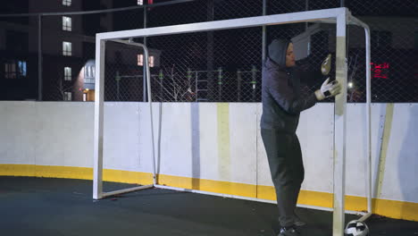 goal keeper exercising with goal post during night training session on urban outdoor sports court, scene includes focus on preparation, illuminated residential buildings in background