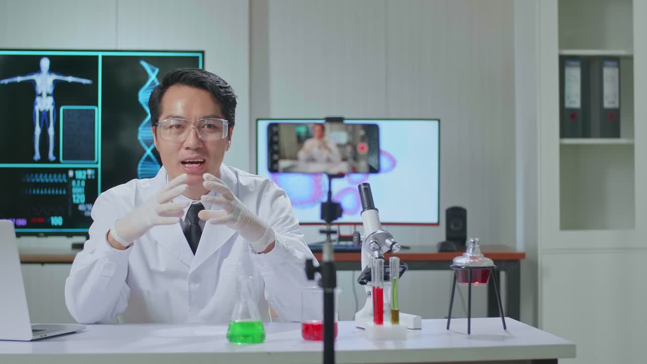 Asian Scientist Man Recording Webinar, Using Smartphone On Tripod, Sitting At Desk In Office