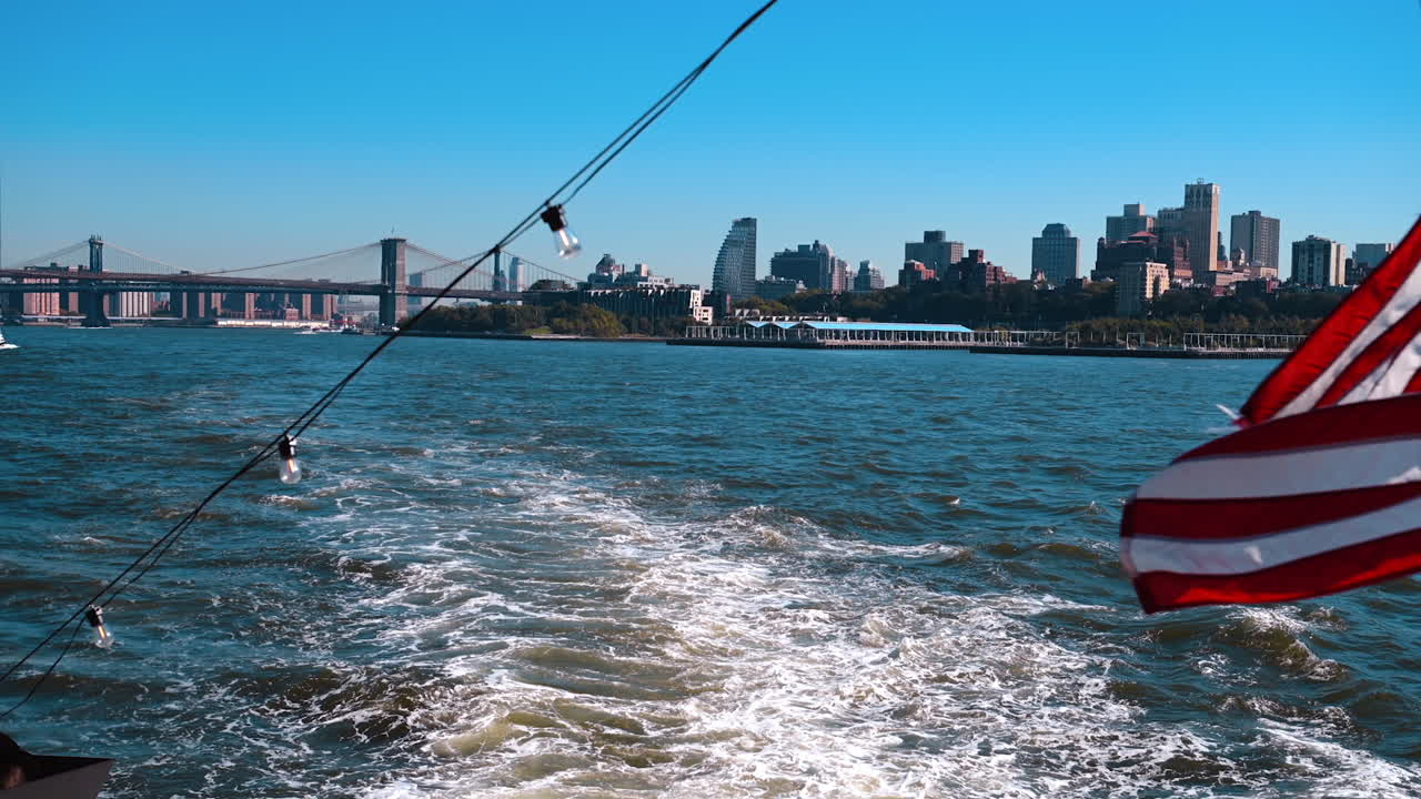 Flag and light bulbs on the boat moving by the river. View on the Brooklyn Bridge and Manhattan Bridge and city skyline at backdrop